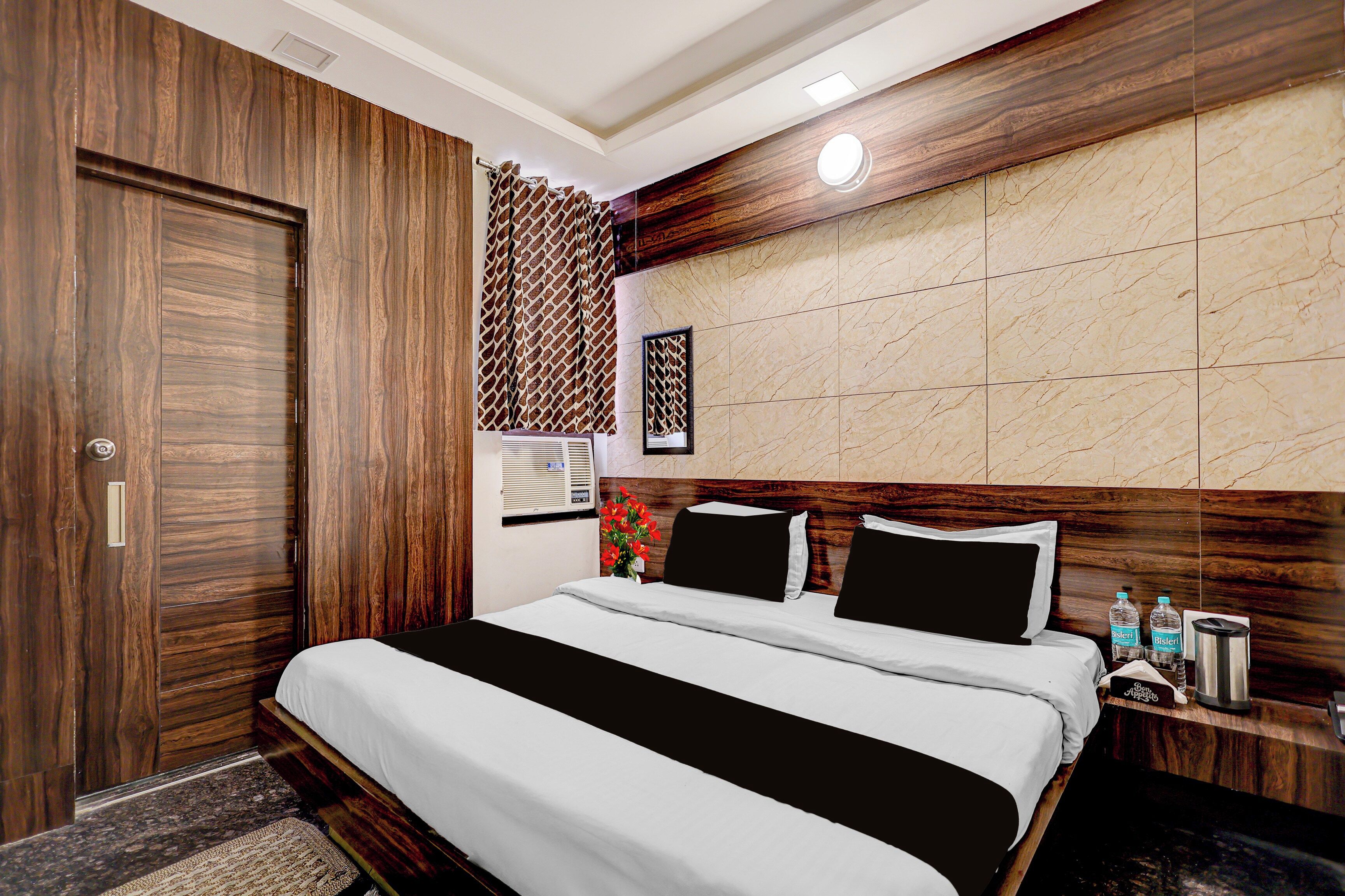 Habitación de hotel con armario de madera, cama con ropa blanca, cabecera con panel que imita el mármol y mesita de noche con una botella de agua y una cafetera.