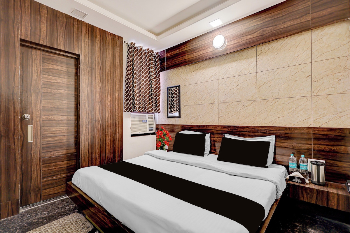A hotel room with a wooden wardrobe, a bed with white bedding, a headboard with a marble-like panel, and a nightstand with a water bottle and a coffee maker.