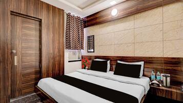 Habitación de hotel con armario de madera, cama con ropa blanca, cabecera con panel que imita el mármol y mesita de noche con una botella de agua y una cafetera.