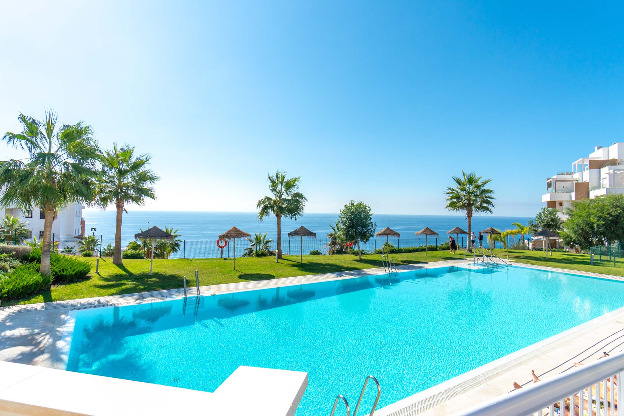 Una piscina blava i cristal·lina amb vista al mar, envoltada de palmeres i un edifici modern.