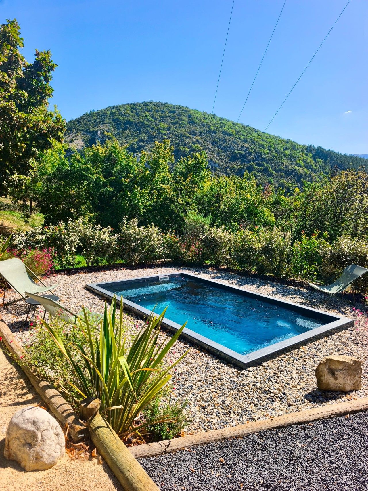 Piscina retangular com superfície de água azul cristalina, rodeada por uma área de cascalho com duas espreguiçadeiras e diversos tipos de plantas.