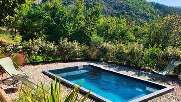 Piscina retangular com superfície de água azul cristalina, rodeada por uma área de cascalho com duas espreguiçadeiras e diversos tipos de plantas.