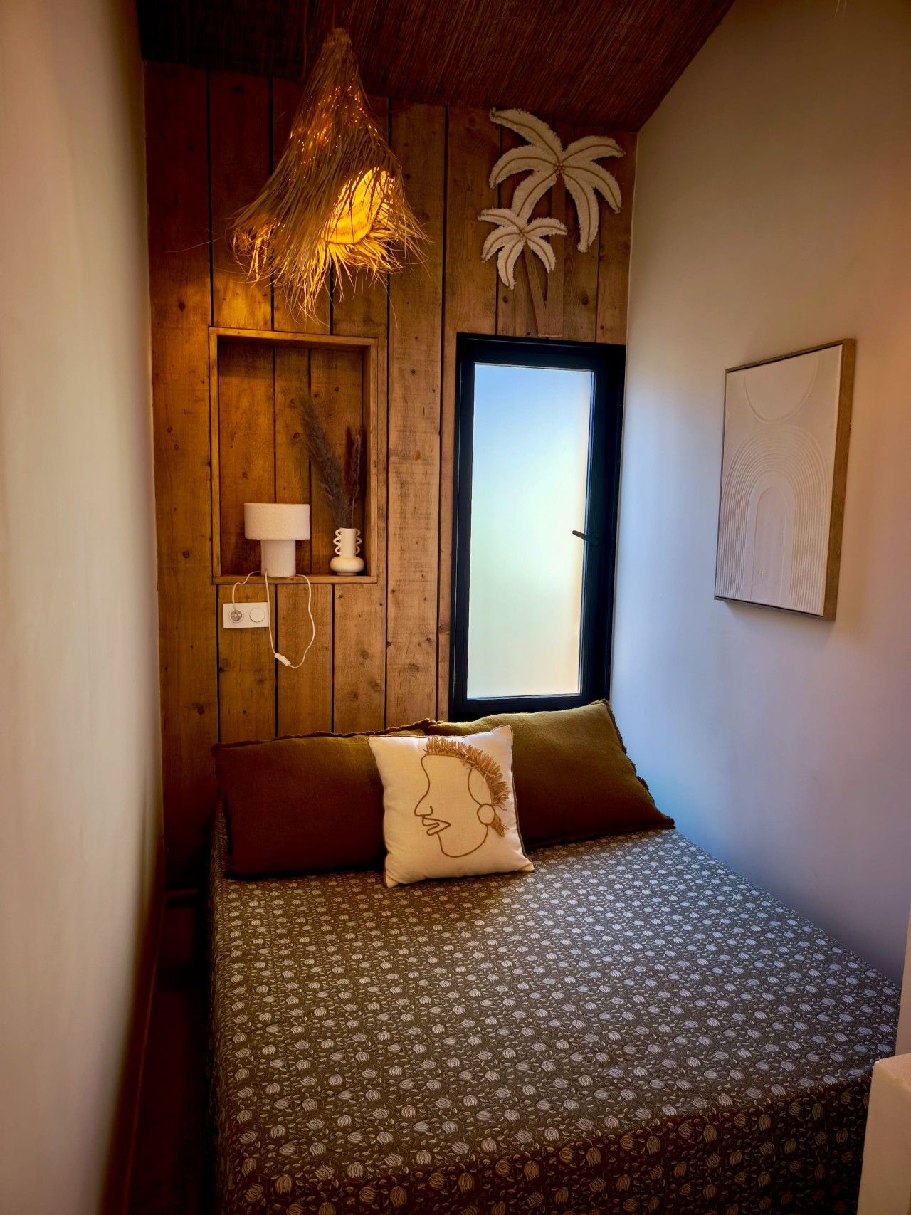 Une chambre à coucher comprenant un lit, une fenêtre, une étagère fixée au mur avec des objets décoratifs et une œuvre d’art murale.