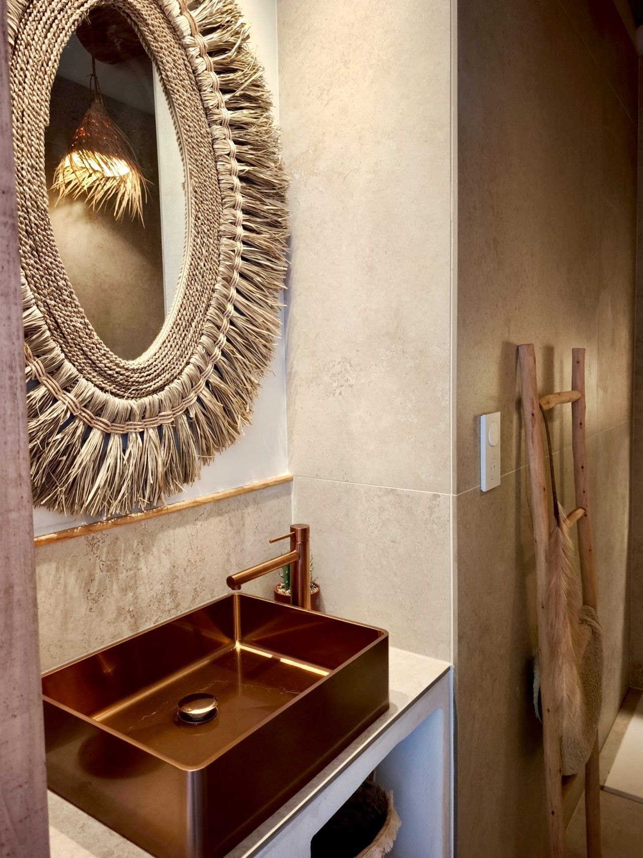 Un baño con espejo tejido, lavamanos de cobre y toallero de madera.