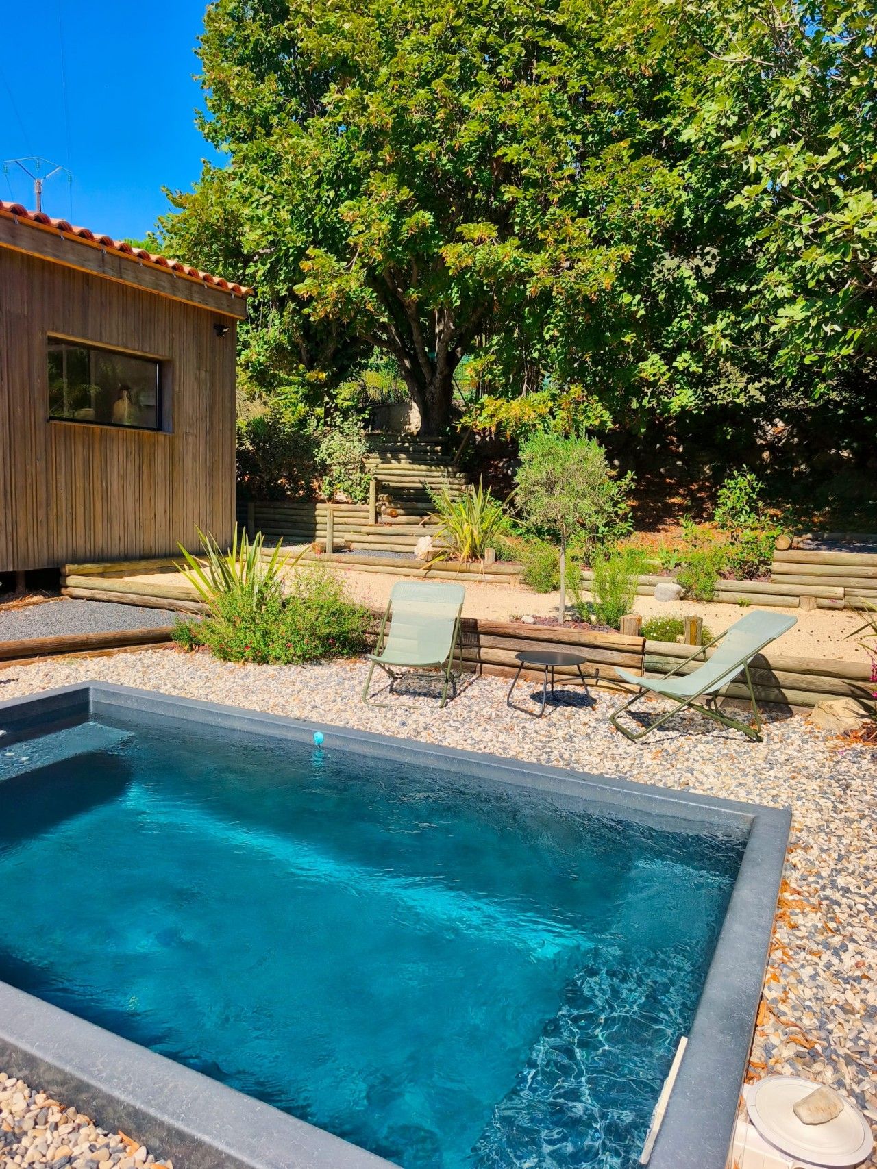 Une piscine avec une zone de terrasse en bois, entourée de galets et de plantes, avec un petit bâtiment et un grand arbre en arrière-plan.