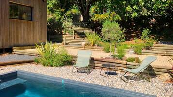 Une piscine avec une zone de terrasse en bois, entourée de galets et de plantes, avec un petit bâtiment et un grand arbre en arrière-plan.