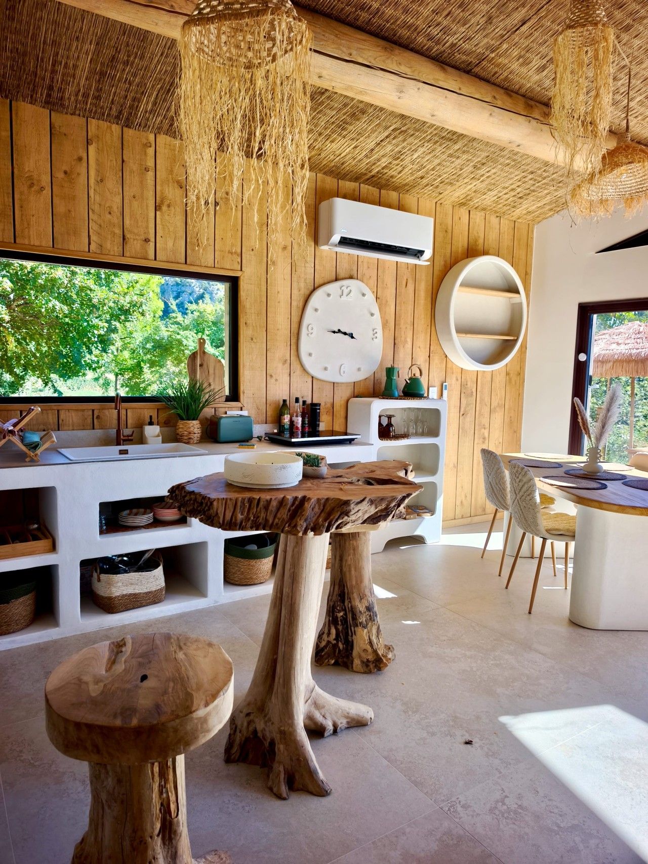 Un intérieur moderne avec un plafond en bois, une grande fenêtre, une horloge murale ronde et un coin repas comprenant une table et des chaises blanches.
