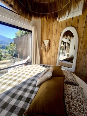 A bedroom with a checkered bedspread, a brown sofa, a hanging macrame wall decoration, and a large mirror reflecting a balcony with a view of the outdoors. - Lodge des Baronnies II - Les Cabanes de Provence (Plaisians)