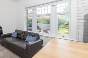 A modern living room with a brown leather sofa, two blue cushions, a wooden table, and large windows offering a view of a garden and a brick building. - Private flat Near Shops & Thames Riverside - Pass the Keys (Reading)