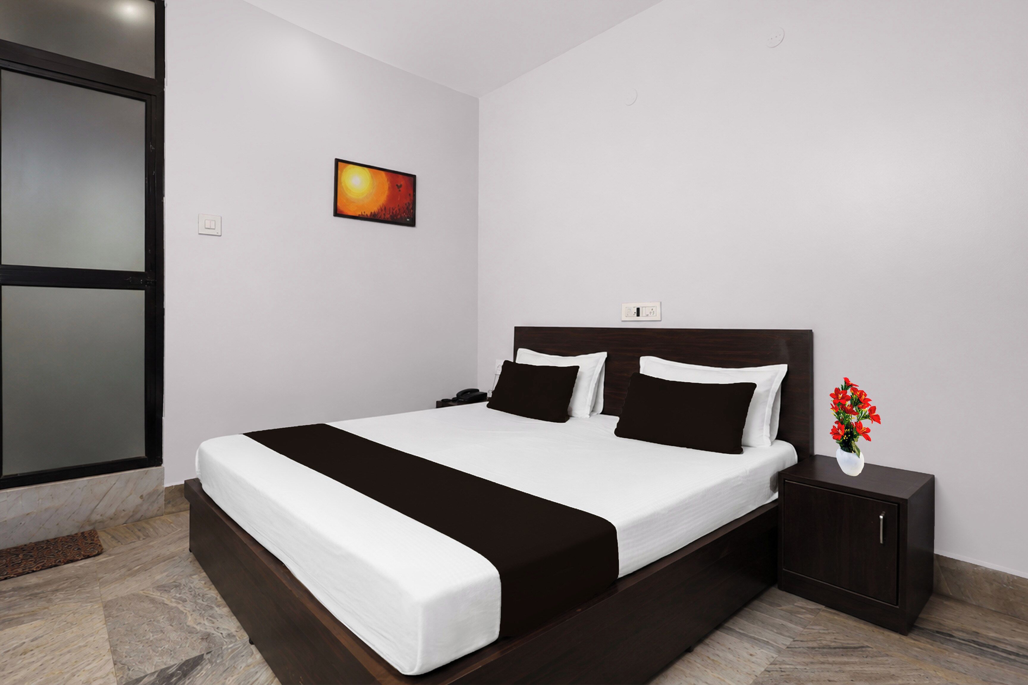 A modern hotel room with a large bed, a nightstand, a vase with flowers, and a painting on the wall.