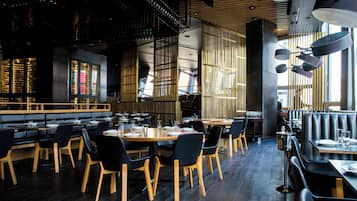 A restaurant interior with wooden tables and chairs, a bar area, and a high ceiling with hanging lights.