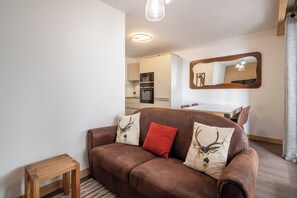 A modern living room with a brown leather sofa, a small wooden table, and a kitchen in the background. - Nanook – Ski-in/Ski-out | Pool | Gym | Alpe d’Huez (Huez)