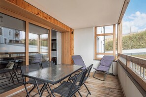 Un espace de restauration extérieur moderne, doté d’une table et de chaises en verre, d’un plancher en bois et de grandes fenêtres offrant une vue sur le paysage environnant.