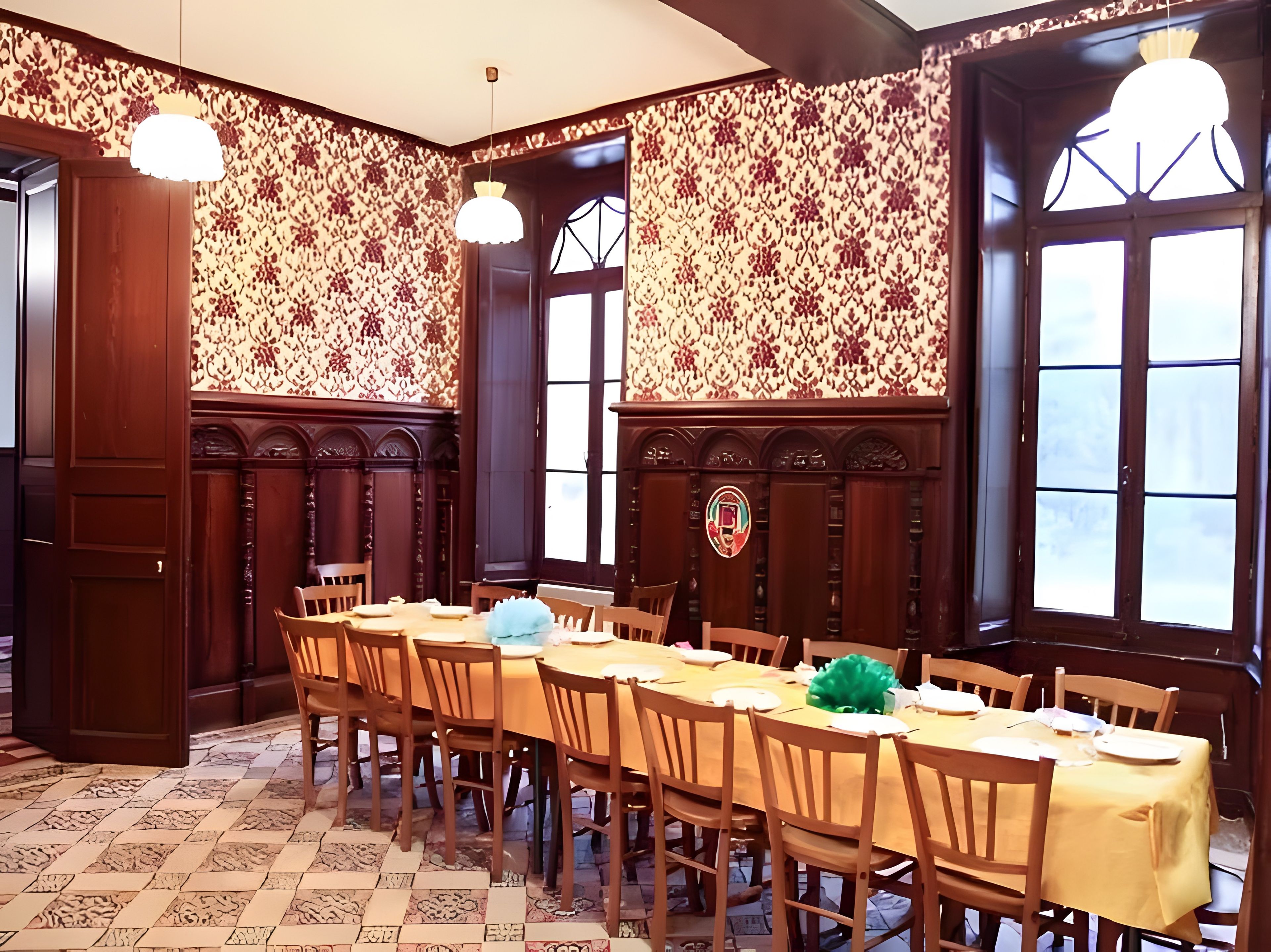 Un espace repas avec une longue table dressée pour accueillir plusieurs personnes, des chaises en bois, un papier peint à motifs et de grandes fenêtres.