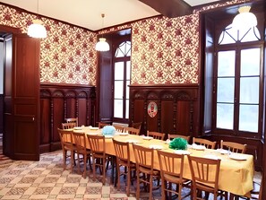 A dining area with a long table set for multiple people, wooden chairs, patterned wallpaper, and large windows.