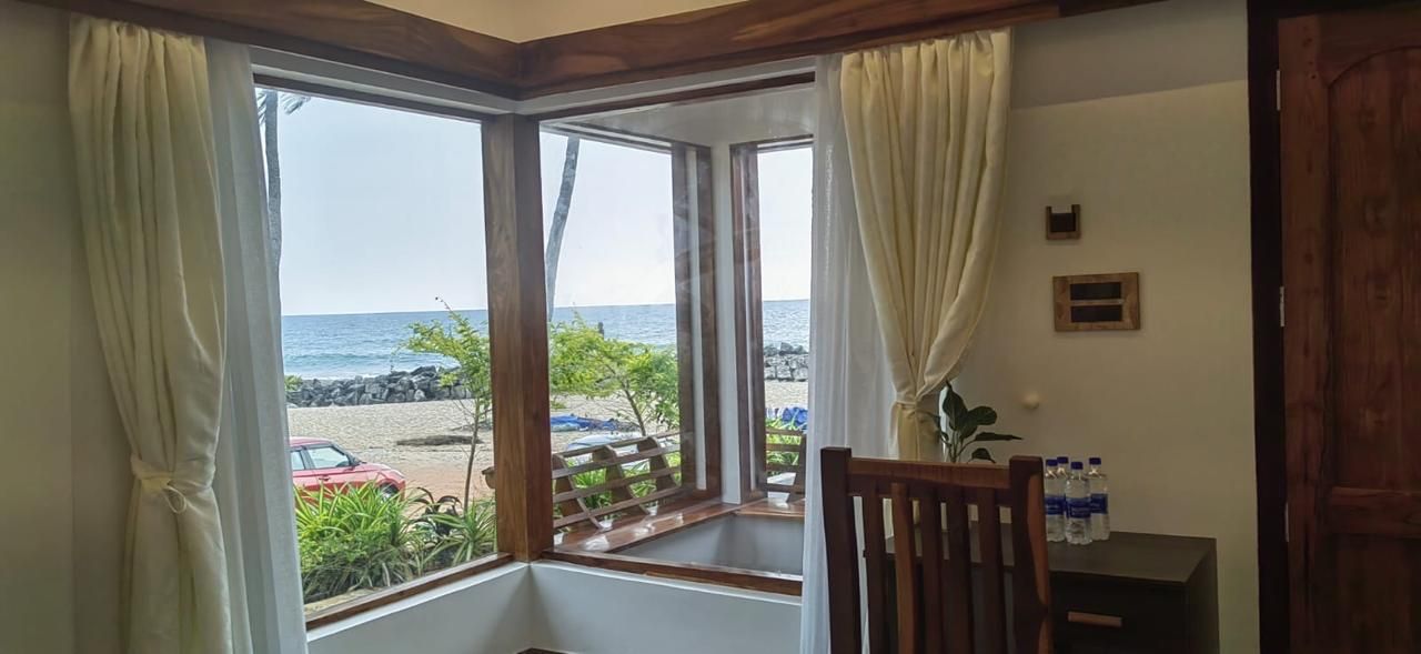 A room with a large window overlooking a beach, a wooden chair, and a small table with bottles.