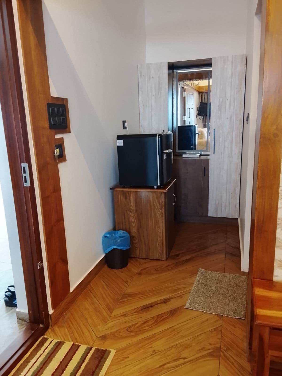 Une pièce avec un plancher en bois, une petite poubelle, un téléviseur posé sur un meuble en bois, et une vue sur un coin cuisine.