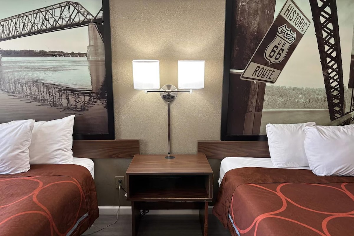 A hotel room with two beds, a nightstand, a lamp, and a mural of a bridge.