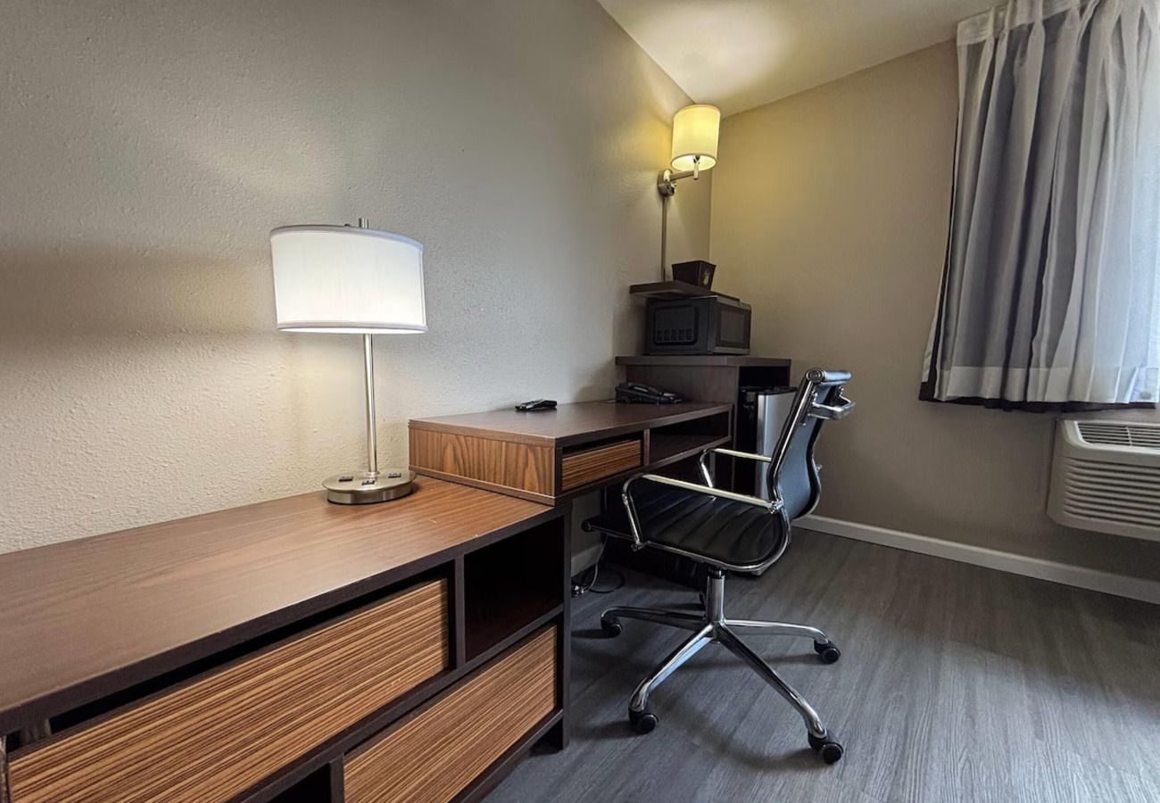 A hotel room with a wooden desk, a black office chair, a lamp, a television, and a window with curtains.