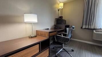 A hotel room with a wooden desk, a black office chair, a lamp, a television, and a window with curtains.