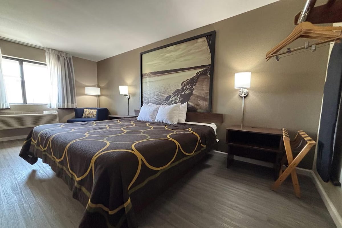A hotel room with a large bed, a bedside table, a chair, a wall-mounted TV, and a window with curtains.