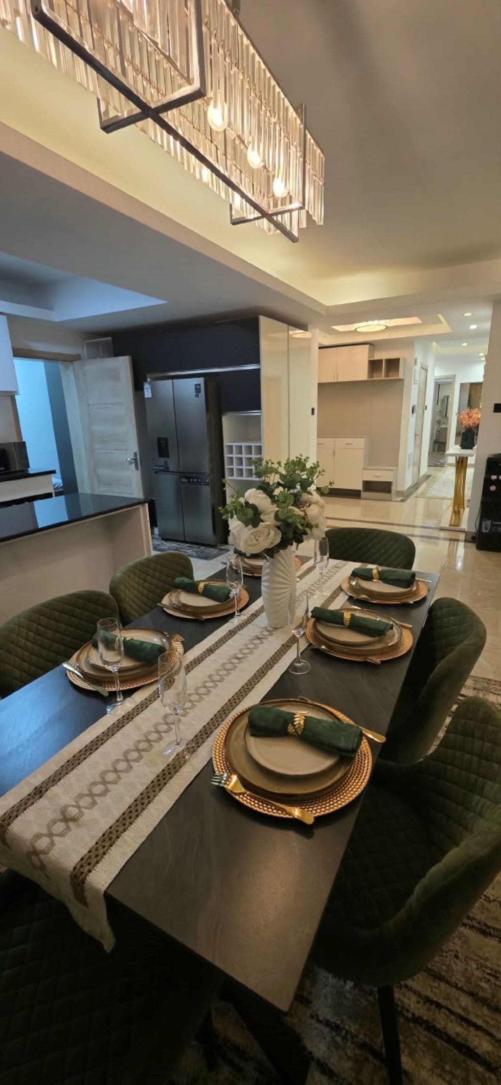 A dining area with a long table set for four, a modern chandelier, and a kitchen in the background.