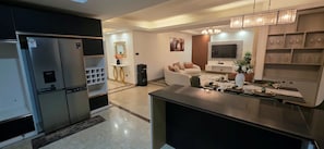 Cozinha moderna com geladeira, área de jantar com mesa posta para dois e sala de estar com sofá e televisão.