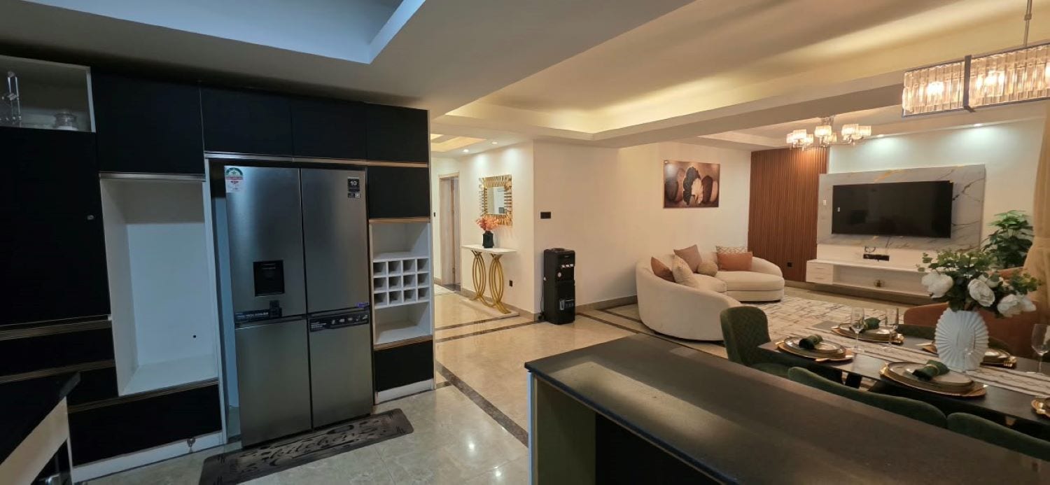 A modern kitchen with a built-in refrigerator, a dining area with a sofa and a TV, and a chandelier.