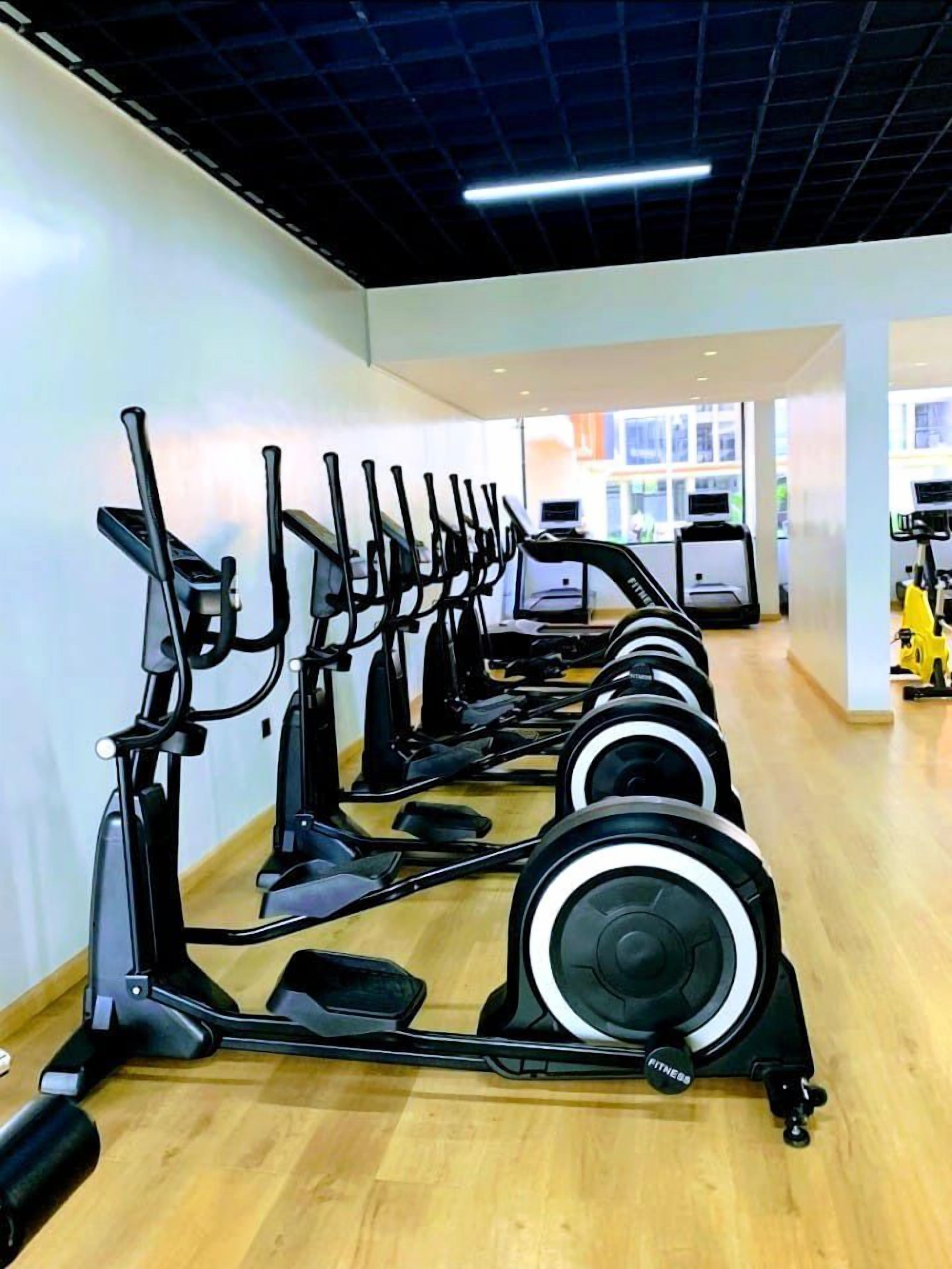 A row of stationary bikes in a gym with a wooden floor and white walls.