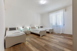 A hotel room with two beds, a wooden floor, a window with curtains, and bedside lamps. - A bright 96-square-meter apartment, perfect for seven people, located on the first floor of a building without an elevator.The property enjoys an ideal location for exploring the streets of the historic center—with its charming vistas, shops, and local (Ferrara)