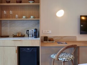 Et moderne køkken med kaffemaskine, elkedel og en kurv med brød. Der er en træstol med et mønstret sæde og en vægmonteret lampe.