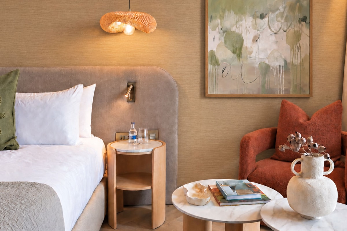 A hotel room with a bed, bedside table, lamp, artwork, and a seating area with a chair and table.