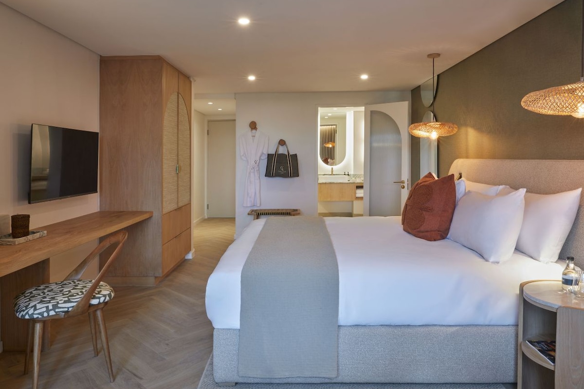 A hotel room with a bed, bedside table, a desk with a chair, a television, and a bathroom visible through an open door.