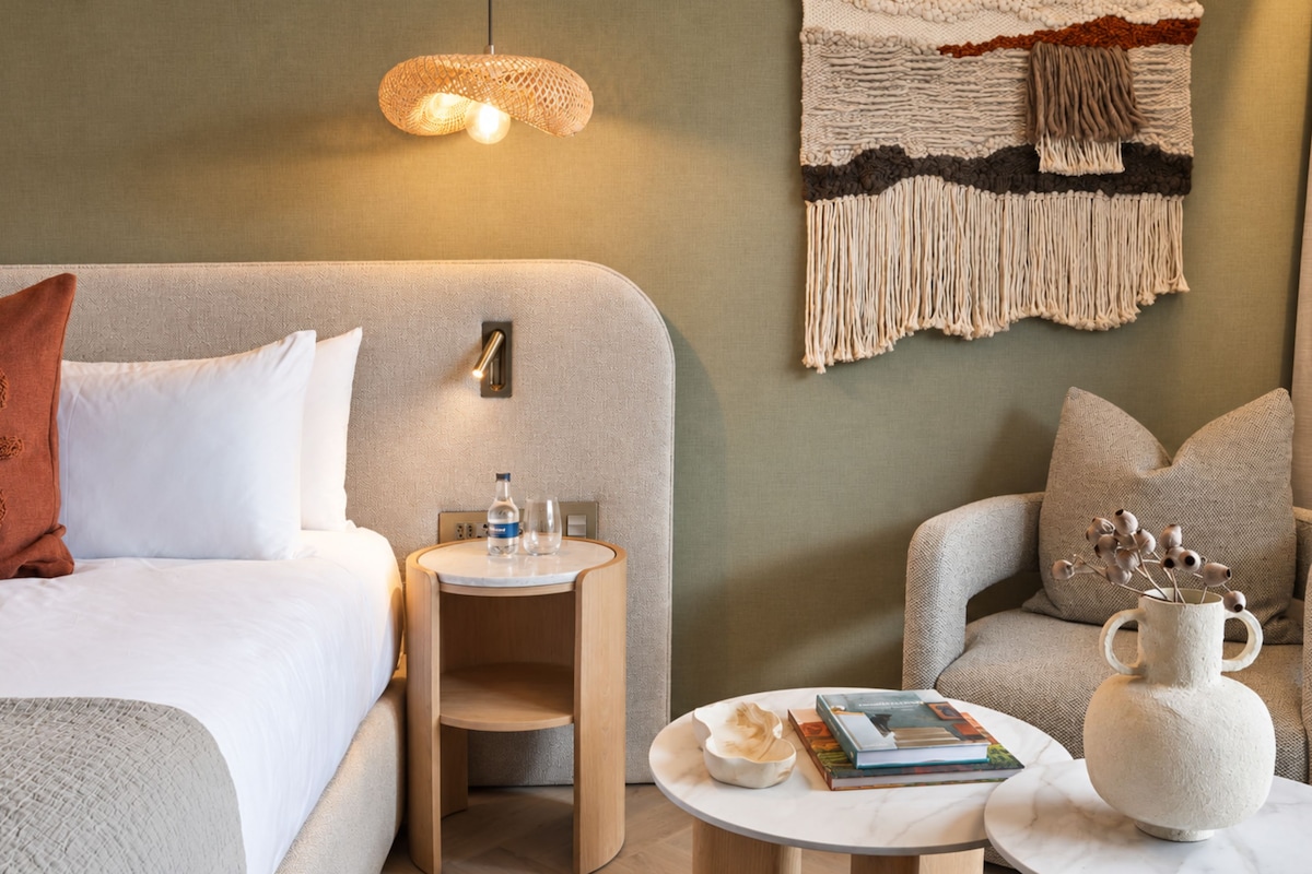 A modern hotel room with a bed, a round wooden side table, a small round table, a wall-mounted lamp, and a decorative wall hanging.