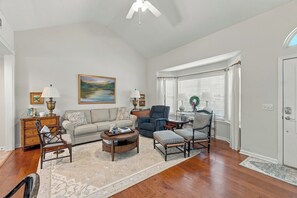 A spacious living room with hardwood flooring, a ceiling fan, a large painting, and a view of the outdoors through a window with white curtains. - Great 3 bedroom 3 bath single story house near Pier/Village and the beach (Saint Simons Island)