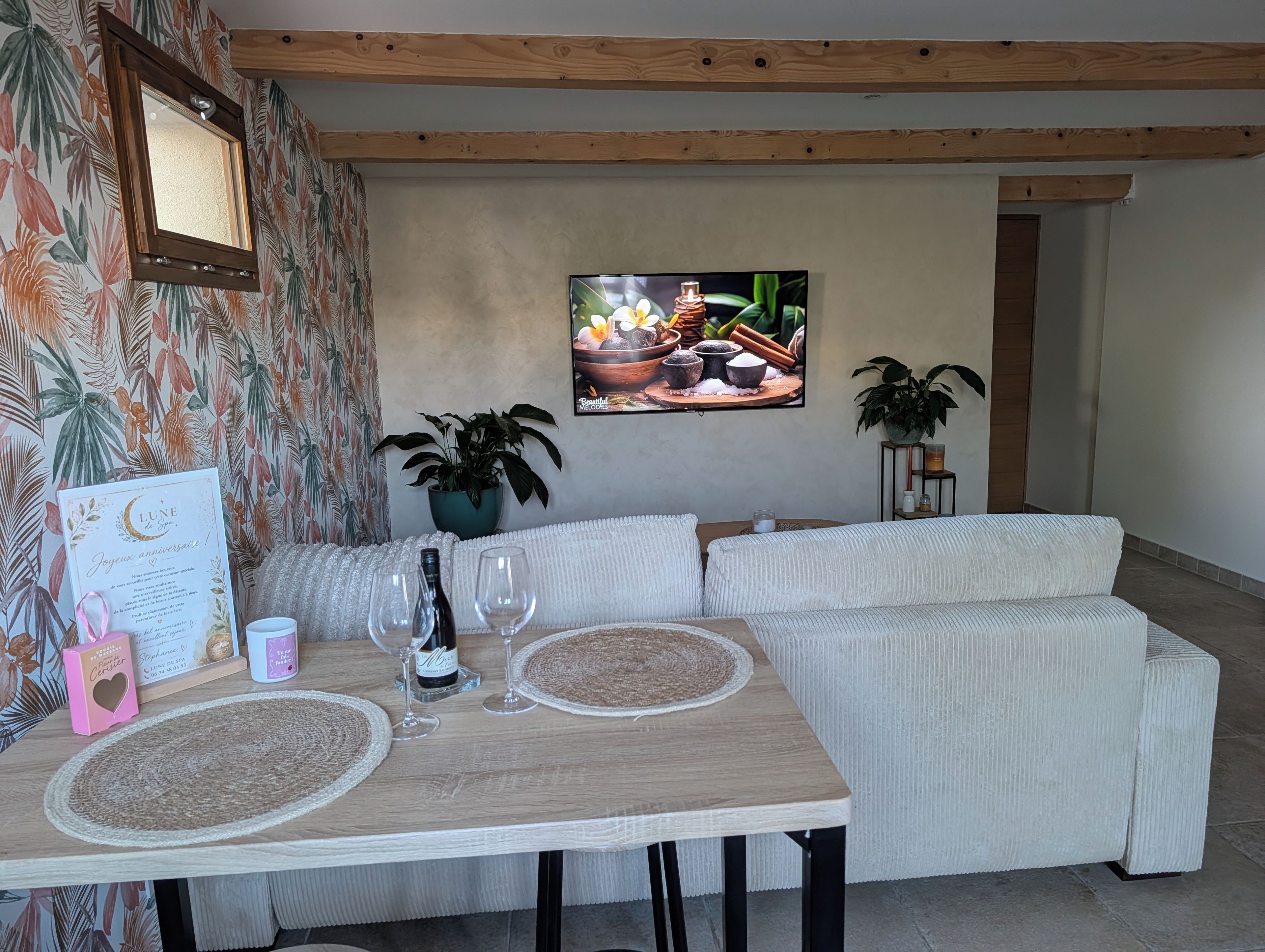 Ein Essbereich mit Tisch für zwei Personen, einer Couch, einer Topfpflanze und einem Fernseher, der eine Kochszene zeigt.