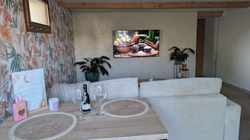Ein Essbereich mit Tisch für zwei Personen, einer Couch, einer Topfpflanze und einem Fernseher, der eine Kochszene zeigt.