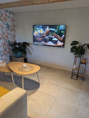 Un salon avec un téléviseur fixé au mur, une table ronde en bois surmontée d’un set de table en fibres tressées, deux plantes en pot et une petite étagère avec des bougies et un récipient.