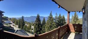 A balcony with a view of a mountain range, a road, and a building with a distinctive roof structure. - A201 - 57 m² - 3-room apartment - 6 people (Prapoutel)