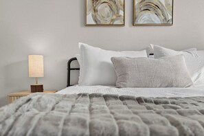 Een net opgemaakt bed met een gebreide deken, een bedlampje en twee abstracte schilderijen aan de muur.