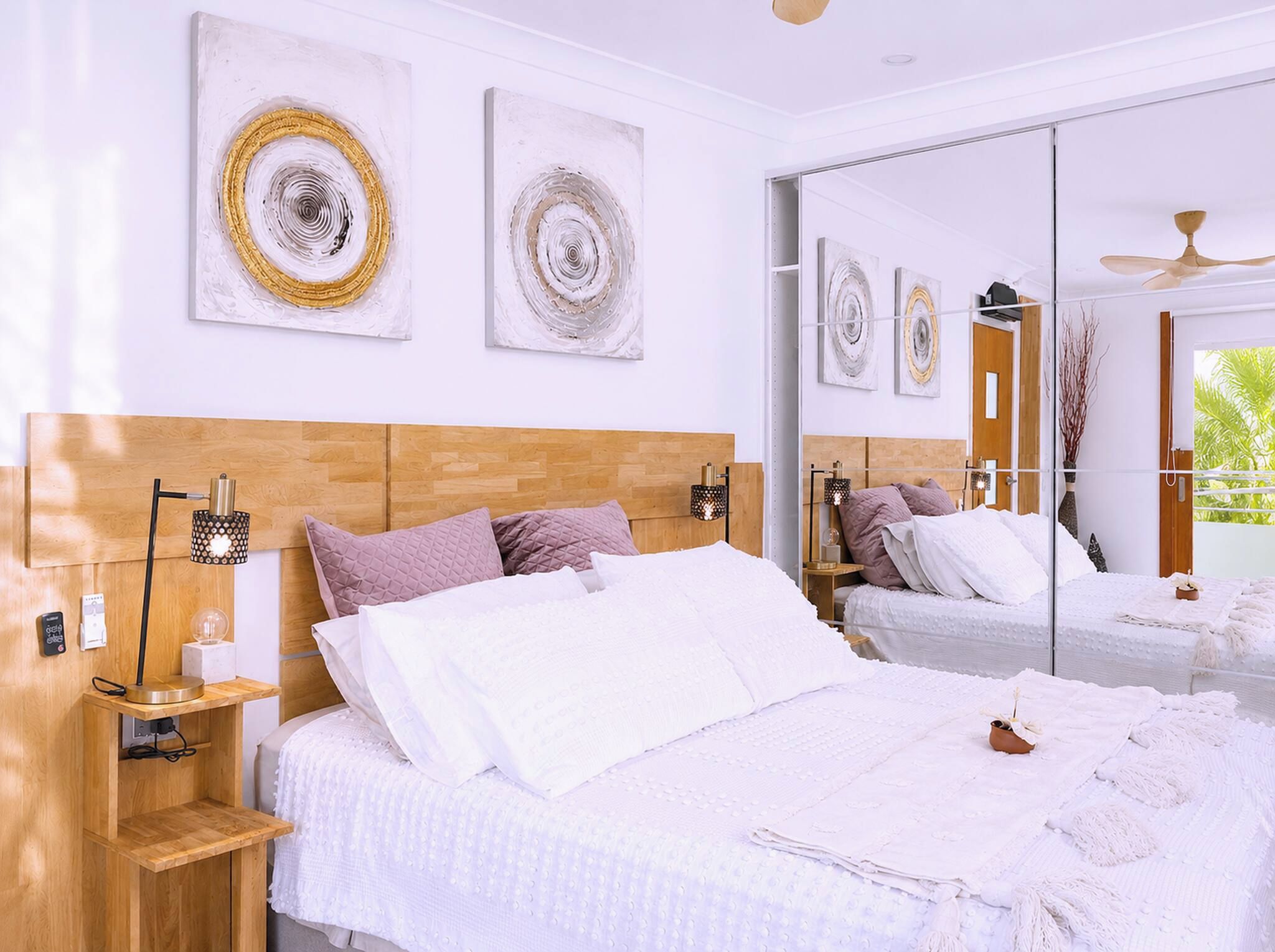 A modern bedroom with a wooden headboard, a large bed with white bedding, a bedside table with a lamp, and a mirror.