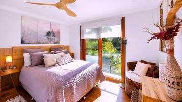 Un dormitorio con cama, mesita de noche, lámpara, ventilador de techo, ventana con vistas a vegetación y un jarrón decorativo.