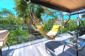 Un balcón con una silla hamaca, una mesa de vidrio con adornos, y vistas de vegetación tropical.
