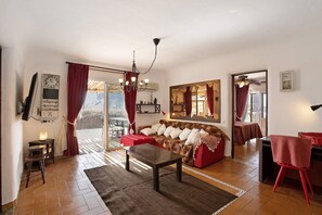 A living room with a sofa, a coffee table, a dining table, and a chair. There is a window with curtains, a wall-mounted clock, and a mirror. - Country House 'Finca Fani 1 con Estudio' with Sea and Mountain Views, Pool, Wi-Fi, Terrace and Garden (Los Corralillos)