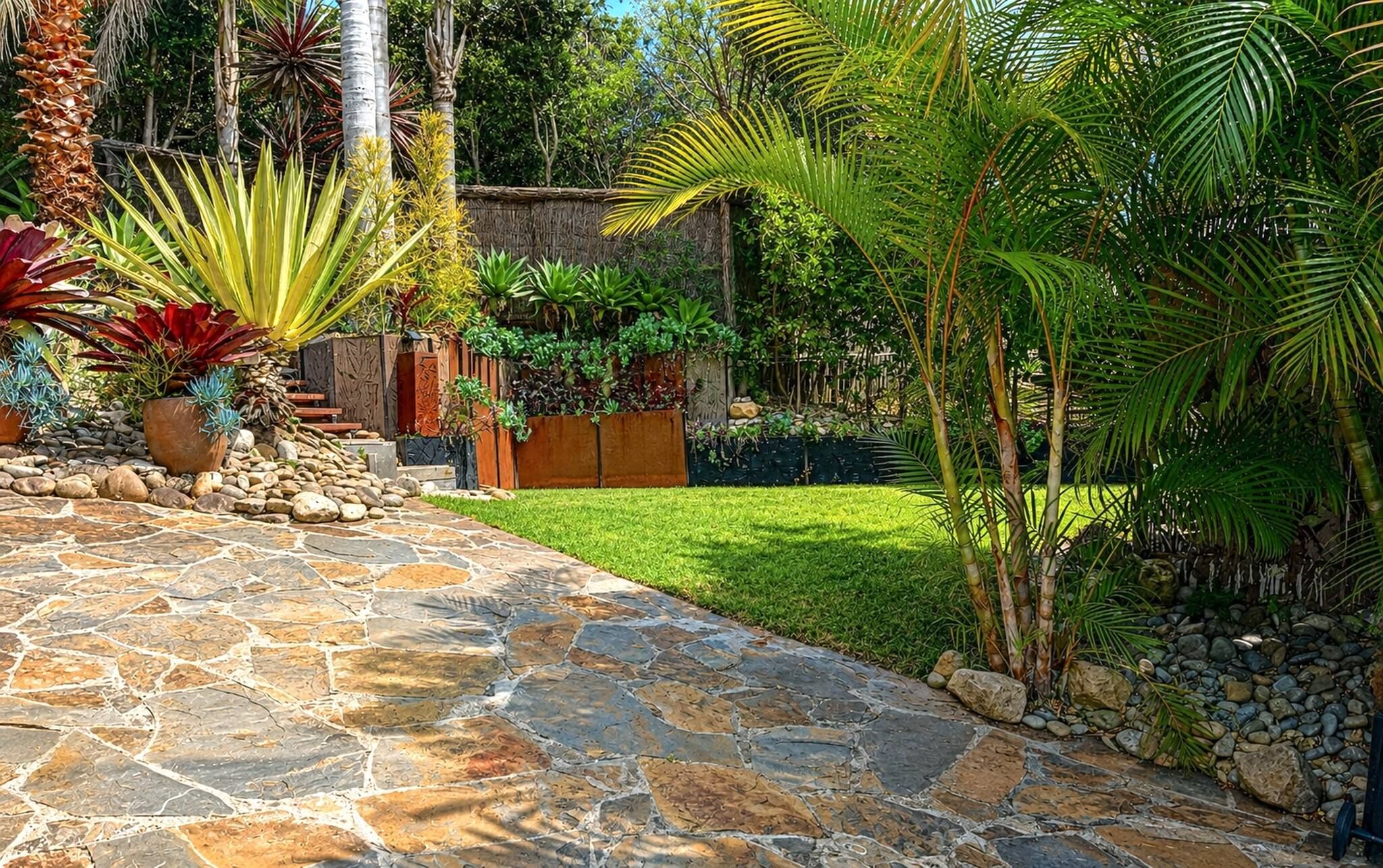 A stone pathway leading to a wooden deck surrounded by lush tropical plants and a well-maintained lawn.