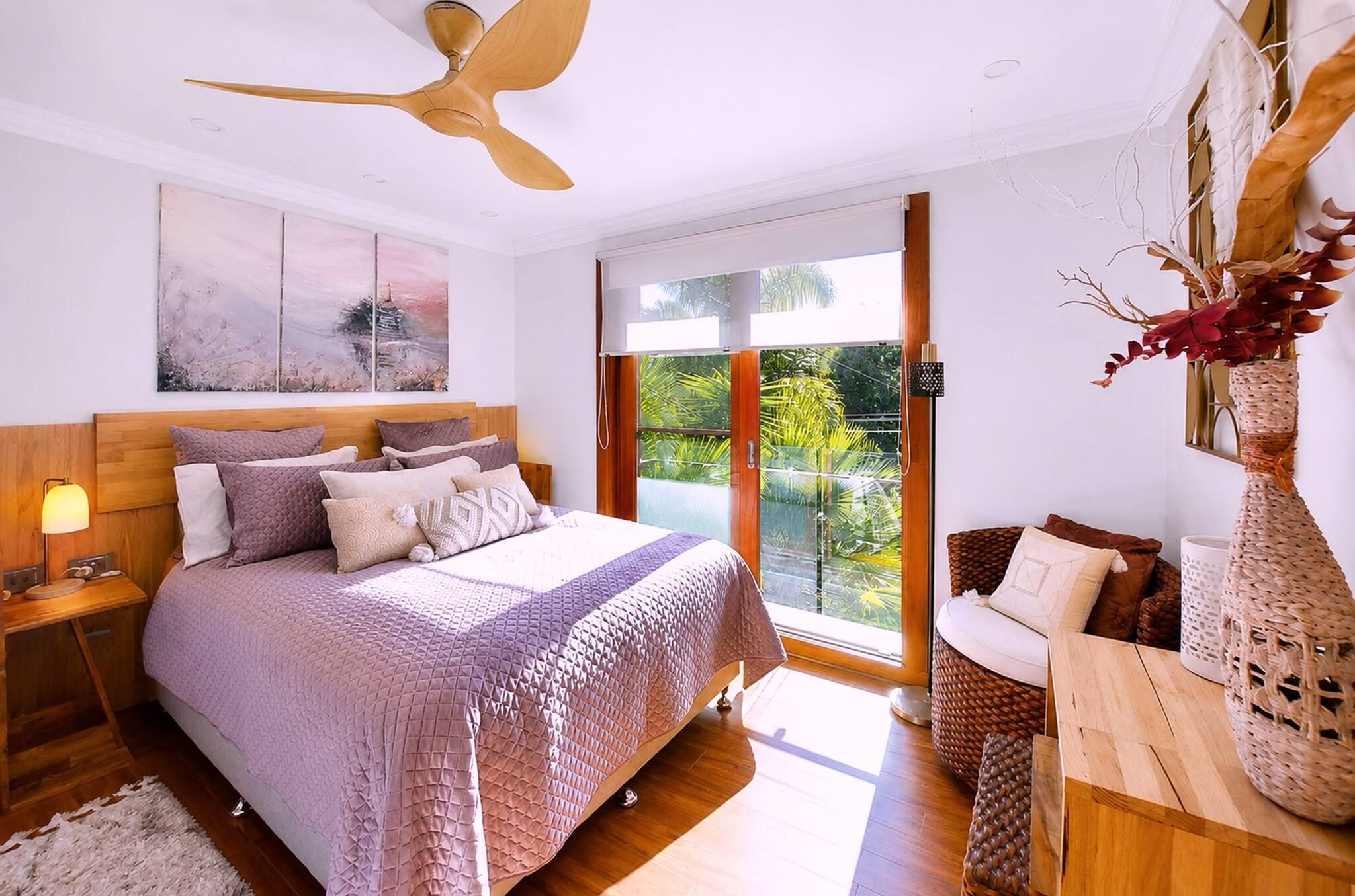 Une chambre à coucher comprenant un lit, une table de chevet, une lampe, un ventilateur de plafond, des œuvres d’art et une vue sur la verdure, visible à travers une porte coulissante.