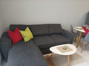 A grey sofa with red and yellow cushions, a small white table with a candle, and a dining area with a red tablecloth. - One Bedroom Apartment 37- Trakia 54 А (Plovdiv)