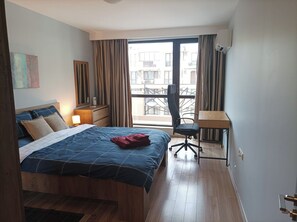 A bedroom with a bed, a desk, a chair, a window with curtains, and a view of buildings outside. - One Bedroom Apartment 37- Trakia 54 А (Plovdiv)