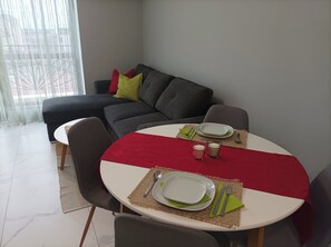 A dining area with a round table set for two, a grey sofa, and a window with sheer curtains. - One Bedroom Apartment 37- Trakia 54 А (Plovdiv)