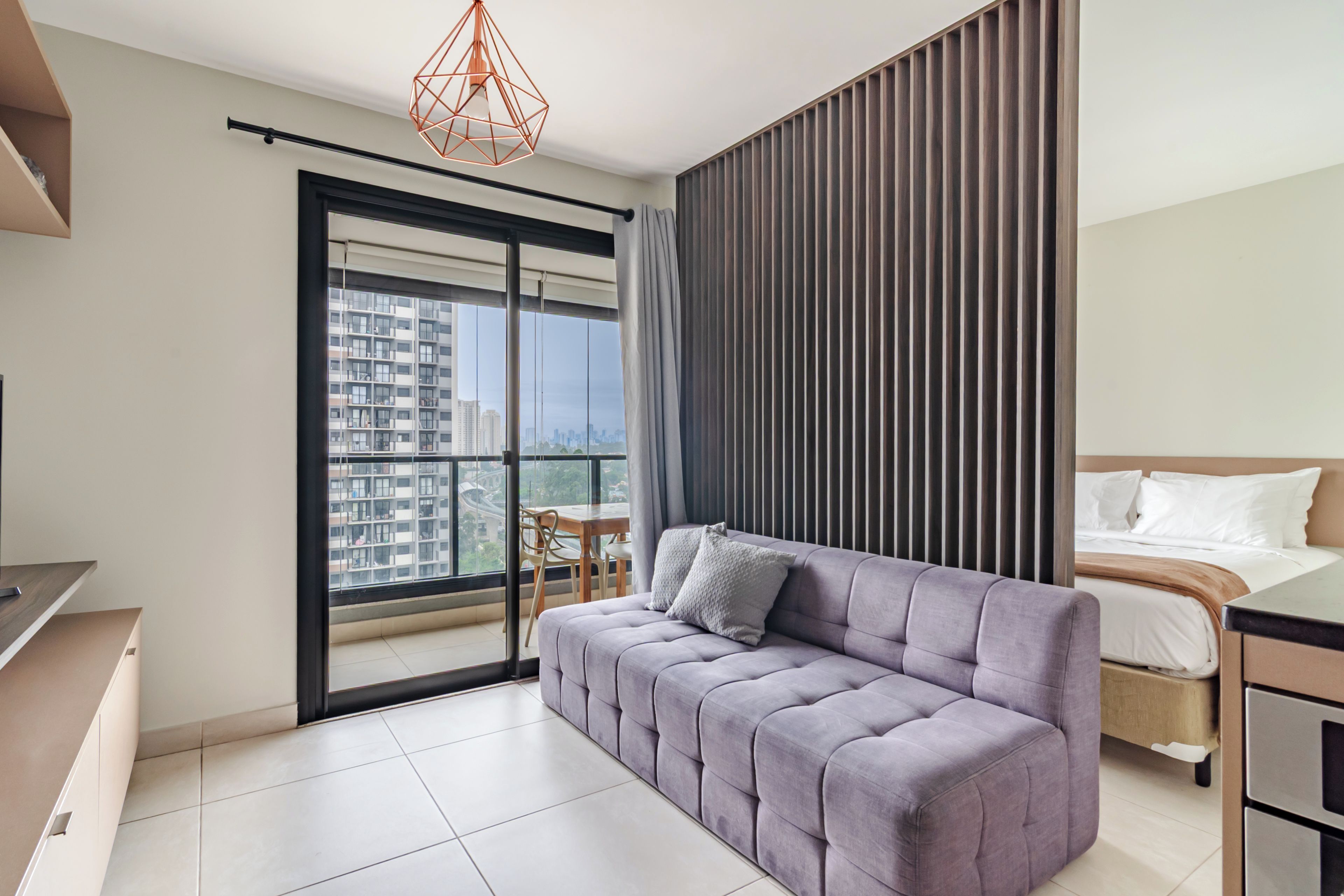 A modern bedroom with a sofa, a bed, and a view of a cityscape through a large window.